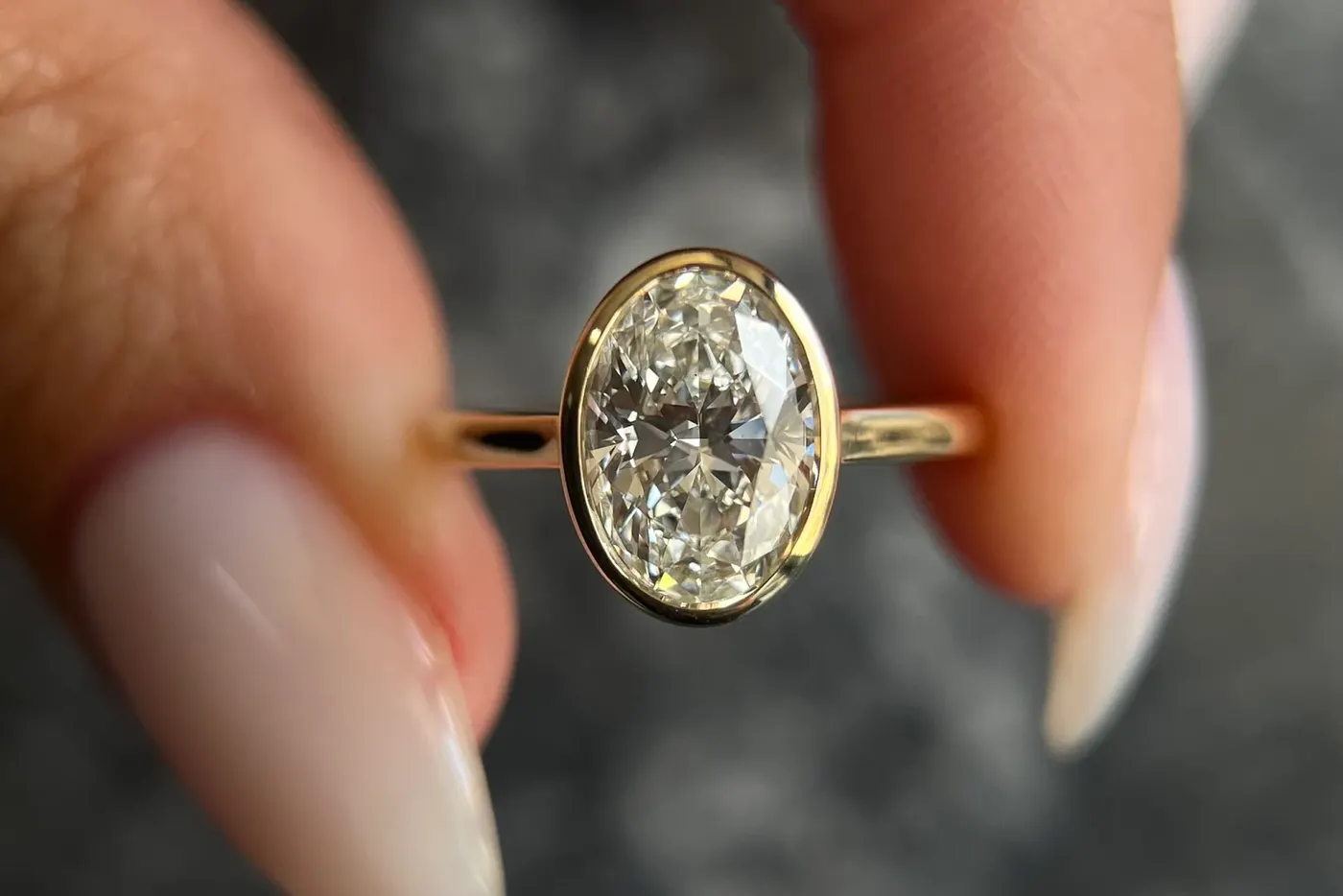 An oval diamond held in a yellow gold bezel setting with no claws, held between finger and thumb