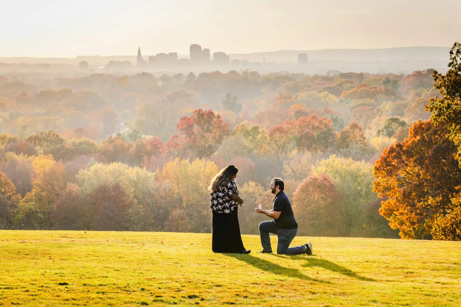 A man kneeling to propose on a quiet hilltop at golden hour, autumn trees and a distant skyline behind